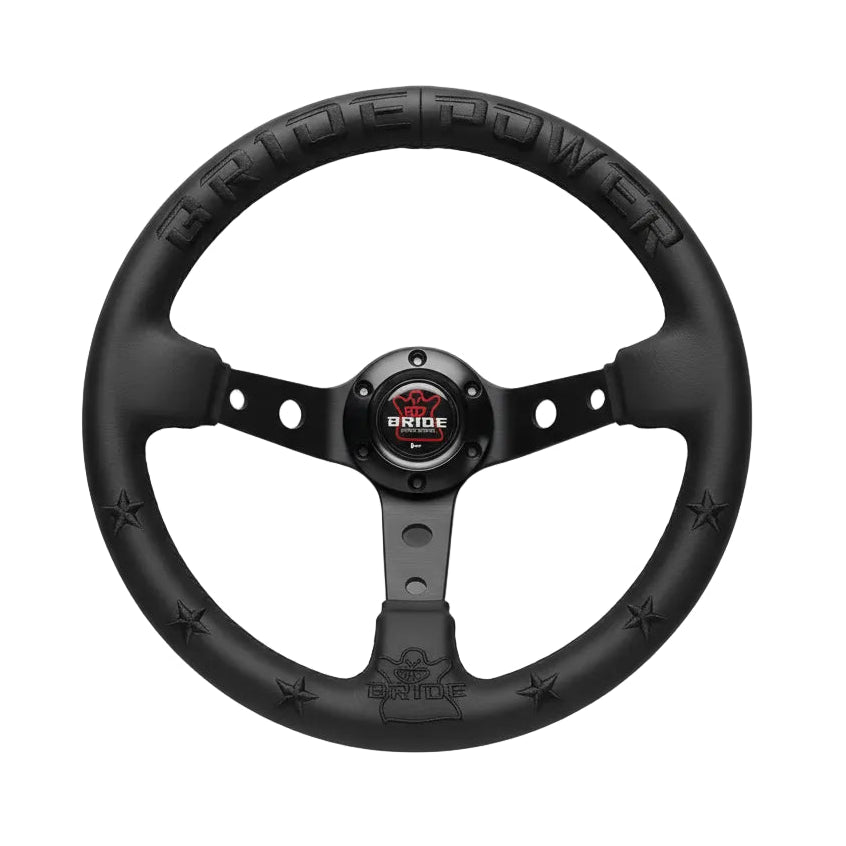 A black Bride steering wheel held by a hand, featuring a matte finish, star accents, and a central logo badge horn button.