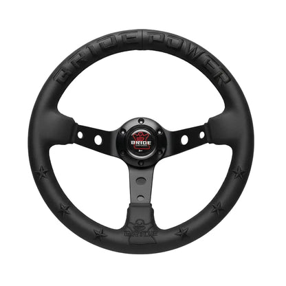 A black Bride steering wheel held by a hand, featuring a matte finish, star accents, and a central logo badge horn button.