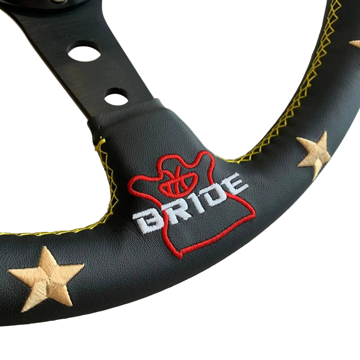 Close-up of a black steering wheel cover with yellow contrast stitching, embroidered red and white 'BRIDE' lettering, and two beige star appliqués on a light wood surface.