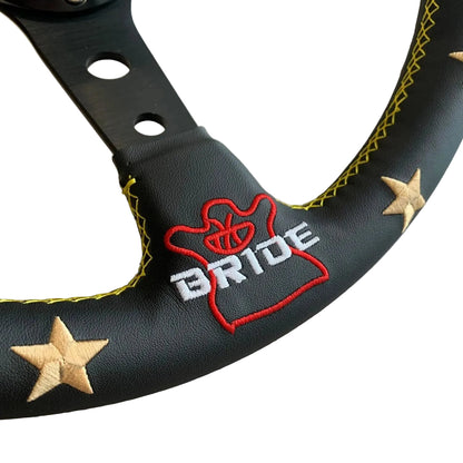 Close-up of a black steering wheel cover with yellow contrast stitching, embroidered red and white 'BRIDE' lettering, and two beige star appliqués on a light wood surface.