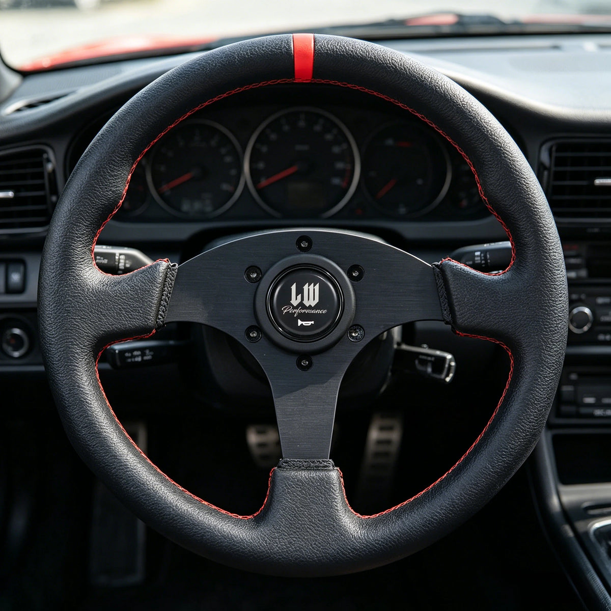 A close-up of a black leather steering wheel with red contrast stitching and a red stripe near the top, featuring a black three-spoke center with a circular logo. The steering wheel is installed inside a car, with the dashboard, instrument cluster, and side air vents visible in the background.