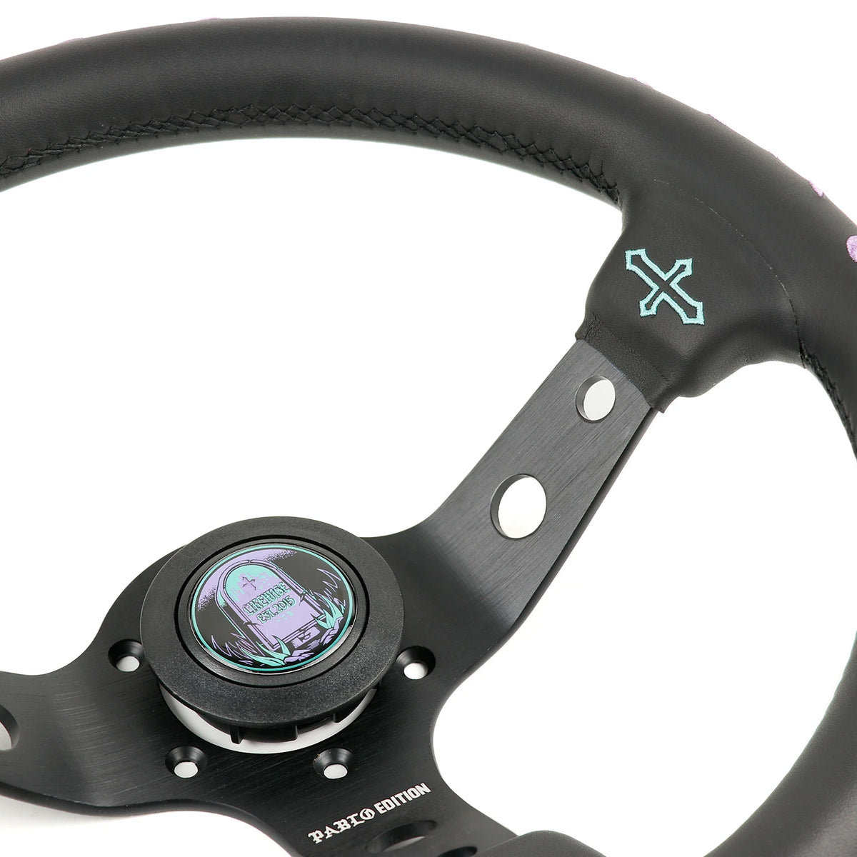 Close-up of a black steering wheel with a brushed metal center hub, a circular display showing a purple map, and a mint green crossed logo on the upper right rim.