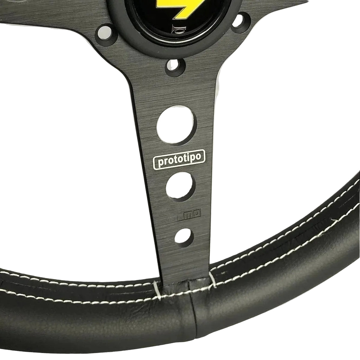 Close-up of a black racing steering wheel with white contrast stitching and a black center cap featuring a yellow arrow logo and 'momo' branding. The center support shows the 'prototipo' brand name.