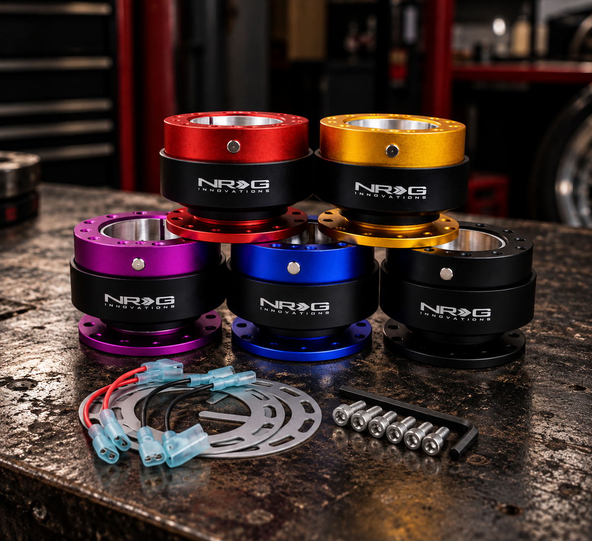 Six NRG innovations steering wheel wheel hubs in red, gold, purple, blue, and black anodized finishes, displayed with mounting hardware on a dark, textured workbench.